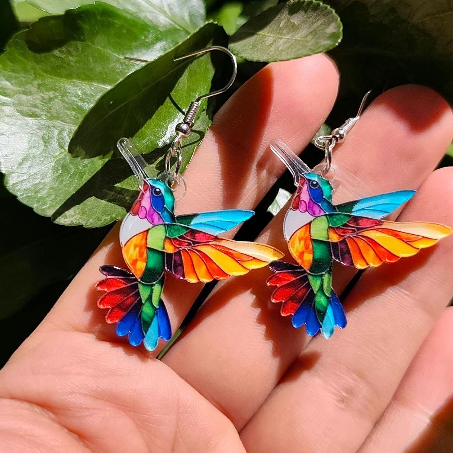Colorful hummingbird-shaped earrings held between fingers with green leaves in the background